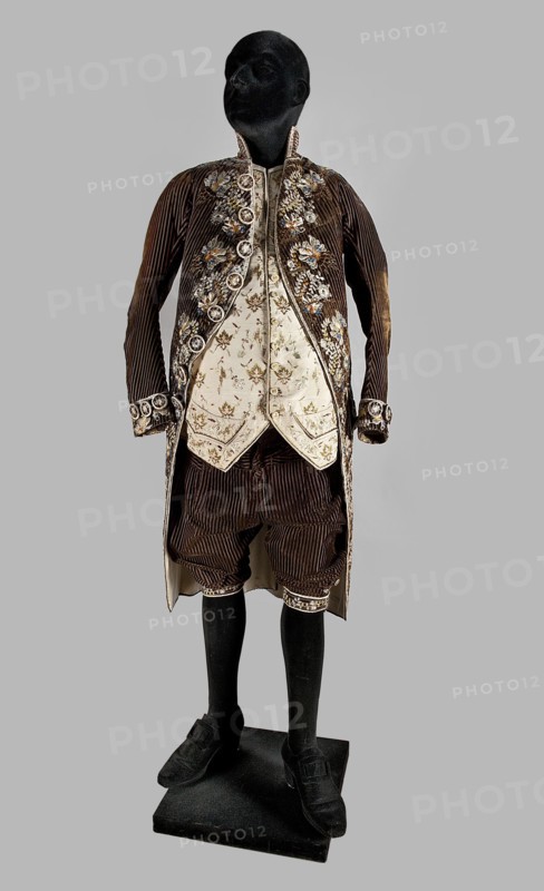 Trousers, trousers, clothing, floral ornaments, men's clothing, men's fashion, 18th century clothing, Trousers, short to the knee, with a lapel, made of striped brown velvet. In the waist, sewn into a wide belt of impregnated brown canvas. The lapel fastens with three covered buttons. The legs have slits, fastened with five buttons each one and a half centimeters in diameter, and finished with narrow, embroidered cuffs. Additional metal and covered buttons for suspenders are sewn on the belt. At the back, below the canvas belt, a triangular gusset made of dark brown smooth velvet is sewn on. 1780s, Silesia, garment, length 76 cm, A mannequin with a black head and black lower legs wears an ornate historical outfit consisting of a dark brown, ribbed long coat with decorative trim and a few floral embellishments, a cream-colored patterned waistcoat beneath with floral motifs, matching cream-colored trim around the coat edges, dark brown knee-length breeches with ribbed texture, black stoc...