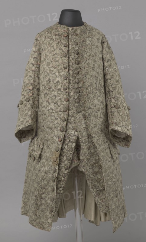 Szustokor, Szustokor made of silver lame with a net ornament featuring a pattern of small flowers. Fitted at the top, widened at the back from hip level down on the sides by folded pleats. Fastened at the front to the neck with seventeen buttons, without a collar. The lapels have curved edges. Long sleeves, set in at elbow height, cut in a curved manner, with wide, turned-up cuffs. The back is cut with a center seam, slit from the hip line, and the flaps do not overlap, decorated with silver braiding at the hem. Fabric-covered buttons are decoratively sewn on the cuffs and under the pocket flaps, four each, and two at the top of the side pleats. Men's garment, 1750–1775, Silesia. Length 108 cm., A long ornate coat displayed on a black mannequin neck, patterned all over with a floral brocade in muted green and beige tones with hints of pale pink, featuring a double-breasted front with a row of fabric-covered buttons, wide sleeves with buttoned cuffs, decorative buttoned flaps near the h...