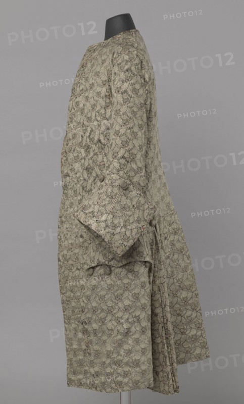 Szustokor, Szustokor made of silver lamé with a net ornament featuring a pattern of small flowers. Fitted at the top, widened at the back from hip level down on the sides by folded pleats. Fastened at the front to the neck with seventeen buttons, without a collar. The lapels have curved edges. Long sleeves, set in at elbow height, cut in a curved manner, with wide, turned-up cuffs. The back is cut with a center seam, slit from the hip line, and the flaps do not overlap, decorated with silver braiding at the hem. Fabric-covered buttons are decoratively sewn on the cuffs and under the pocket flaps, four each, and two at the top of the side pleats. 1750–1775. Silesia. Garment; length 108 cm., A side view of a long patterned coat displayed on a black mannequin head against a neutral gray background, the coat showing a repeated floral-like allover motif in muted green with accents of darker green and subtle brown, a few rounded fabric-covered buttons on the cuff and near the waist, a folded...
