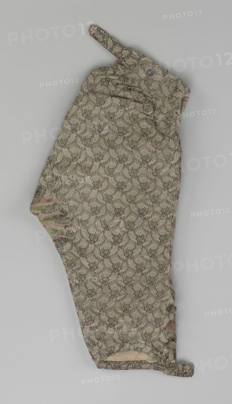 Trousers, short knee-length trousers sewn into a wide belt with a buttoned fly, made of silver lamé with a net ornament featuring a pattern of small flowers. Slit pockets on the sides of the fastener, fastened with a button on the side. The belt is slit at the back and adjustable with a flap. The legs are fastened with four buttons at the bottom and finished with narrow cuffs. Men's clothing, men's fashion. 1750–1775, Silesia. Length 76 cm., A single textile garment laid flat, showing a patterned fabric in muted beige with repeating floral-like motifs in soft green and faded pink, overlaid on a darker grayish lace-like network; the piece has a narrow tab at one end and a rounded tab at the opposite end, a flap with a round button near one edge, a slotted opening near the other edge, and a row of fabric-covered round buttons along one side; parts of the fabric show wear with frayed edges and small areas where the underlying lighter material and threads are visible, and faint irregular d...