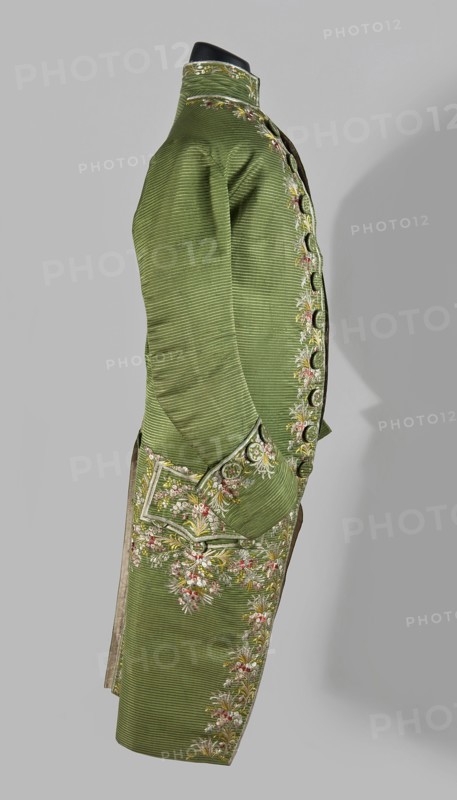 Habit à la française, repossession provenance; floral and plant motifs; embroidery; 18th century clothing. Habit of green silk embroidered with motifs of small bouquets of flowers. Fitted at the top, widened at the back from the waist down with two slit pleats. Fastens at the front with hooks and eyes. Semicircularly cut lapels overlap at breast level and are cut off at the bottom. High standing collar. Long sleeves set in at elbow height, cut in an arc, finished with wide, slightly protruding cuffs. Side pockets at hip level covered with trapezoidal flaps. Back with a center seam. Slits at hip level. Center back flaps meet without a pleat. Embroidery runs in a wide band around the edges, cuffs, pockets, collar, and back pleats. Decorative buttons sewn in threes on the cuffs, under the pocket flaps, two at the top of the back pleats, and in the pressed pleats. 1770s, Silesia place of origin and France place of creation; garment; length 112 cm., A side view of a long green coat displaye...