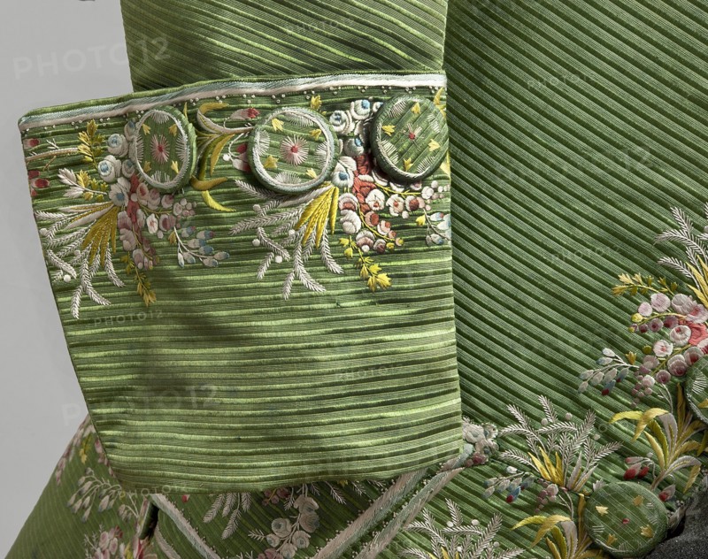 Habit à la française, Repossession provenance. Floral and plant motifs; fashion clothing: men's 18th-century garment. Green silk habit embroidered with small bouquets of flowers. Fitted at the top, widened at the back from the waist down with two slit pleats. Fastens at the front with hooks and eyes. Semicircularly cut lapels overlap at breast level and are cut off at the bottom. High standing collar. Long sleeves set in at elbow height, cut in an arc, finished with wide, slightly protruding cuffs. Side pockets at hip level covered with trapezoidal flaps. Back cut with a center seam; slits at hip level; center back flaps meet without a pleat. Embroidery in a wide band around edges, cuffs, pockets, collar, and back pleats. Decorative buttons sewn in threes on cuffs, under pocket flaps, two at top of back flaps, and in pressed pleats. 1770s, Silesia; France origin/creation. Garment length 112 cm., A close-up of a green textured fabric with diagonal ribbing and a few round fabric-covered ...