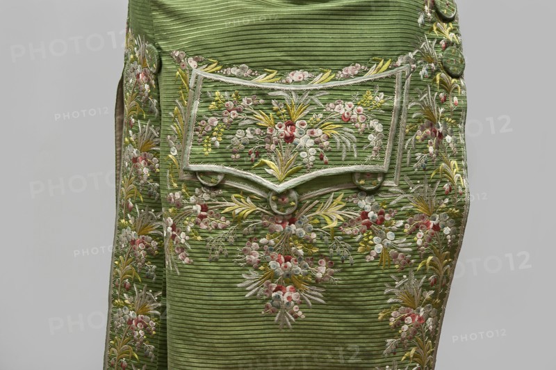 Habit à la française, repossession provenance; floral and plant motifs; fashion, men's clothing; embroidery; 18th century clothing. Green silk habit embroidered with small bouquets of flowers. Fitted at the top, widened at the back from the waist down with two slit pleats. Fastens at the front with hooks and eyes. Semicircularly cut lapels overlap at breast level and are cut off at the bottom. High standing collar. Long sleeves set in at elbow height, cut in an arc, finished with wide slightly protruding cuffs. Side pockets at hip level covered with trapezoidal flaps. Back cut with a center seam; slits at hip level; center back flaps meet without a pleat. Embroidery in a wide band around edges, cuffs, pockets, collar, and back pleats. Decorative buttons sewn in threes on cuffs, under pocket flaps, two at top of back pleats, and in the pressed pleats. 1770s, Silesia; made in France. Garment; length 112 cm., A section of a green ribbed fabric panel decorated with ornate floral embroidery...