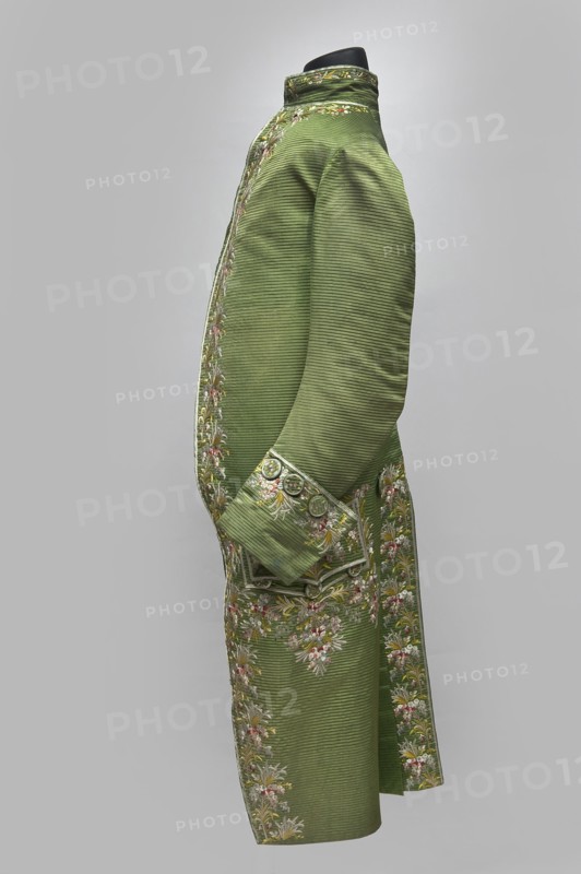 Habit à la française, Repossession provenance. Floral and plant motifs; embroidery. 18th century 1770s. Habit of green silk embroidered with small bouquets of flowers. Fitted at the top, widened at the back from the waist down with two slit pleats. Fastens at the front with hooks and eyes. Semicircularly cut lapels overlap at breast level and are cut off at the bottom. High standing collar. Long sleeves set in at elbow height, cut in an arc, finished with wide slightly protruding cuffs. Side pockets at hip level covered with trapezoidal flaps. Back cut with a center seam; slits at hip level. Center back flaps meet without a pleat. Embroidery runs in a wide band around the edges, cuffs, pockets, collar, and back pleats. Decorative buttons sewn in groups of three on the cuffs, under pocket flaps, two at the top of the back pleats, and in the pressed pleats. Silesia place of origin and France place of creation. Garment; length 112 cm., A side view of a long tailored coat displayed on a bl...