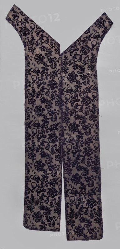 Fabric fragment, textiles, fragments of silk fabric, patterned fabric with plant motifs; cochineal and indigo dyes; fragment of a larger textile object. Patterned, looped purple velvet with plant motifs. Motif: plant tendril of small fanciful floral twigs in asymmetrical arrangements creating a net pattern. Weave construction: grosgrain background with silk bold and linen weft in natural color, purple silk warp; looped silk purple cover. Dense plant composition of finely curved twigs with varied leaves and flowers—delicate, refined, segmented pattern. Arranged in rows with a half-report shift; calyx of dominant flower alternately tilted left or right in the rows. Design repeat report length 24.7 cm × width 13.2 cm. 17th century, Italy. Fabric dimensions: height 110 cm, width 18 cm., A long fabric garment shaped with a deep V neckline and two wide straps, laid flat against a light gray background, showing a repeated ornate floral and scroll pattern in dark purple on a pale grayish-beige...