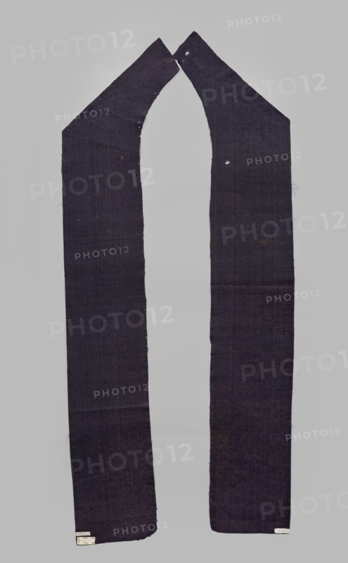Fabric fragment, textiles, fragments of silk fabrics, patterned fabrics, plant motifs, cochineal dye, indigo dye, fragments of a larger textile object, patterned, looped, purple velvet, with plant motifs. Motifs: plant tendril made of small, fanciful floral twigs in asymmetrical arrangements creating a net pattern. Weave construction: grosgrain background with silk and linen weft in a natural color, purple silk warp; looped silk purple cover. Dense plant composition, finely curved twigs with a variety of leaves and flowers – very delicate and with a refined, segmented pattern. Arranged in rows, with a half-report shift and with the calyx of the dominant flower tilted alternately to the left or right in the rows. Design report length 24.7 x width 13.2 cm. Maker/attribution: Justyna Boninska. 17th century, Italy. Fabrics, height 110 cm, width 18 cm., A long, narrow fabric piece with a U-shaped central cutout, shown against a pale gray background; the fabric is a dark indigo or deep navy ...