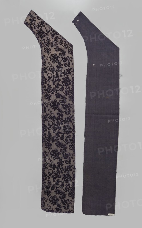 Fabric fragment, fragments of silk patterned fabric, patterned purple looped velvet with plant motifs. Plant tendril made of small fanciful floral twigs in asymmetrical arrangements creating a net pattern. Weave construction: grosgrain background with silk and linen weft in a natural color, purple silk warp; looped silk purple cover. Dense plant composition, finely curved twigs with a variety of leaves and flowers, arranged in rows with a half-report shift and with the calyx of the dominant flower tilted alternately left or right. Design report length 24.7 x width 13.2 cm. Maker: Justyna Boninska, 17th century, Italy. Fabric dimensions: height 110 cm, width 18 cm. Materials/dyes noted: silk, linen; cochineal and indigo dyes., A vertical image shows two long fabric pieces side by side against a pale gray background. The left piece has a beige base with an intricate dark purple floral damask pattern running across its length and a slightly curved upper end. The right piece is a solid dee...