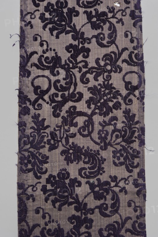 Fabric fragment, textiles, fragments of silk fabrics, patterned fabrics with plant motifs, cochineal and indigo dyes; fragments of a larger textile object. Patterned, looped purple velvet with plant motifs. Report motifs: plant tendril made of small, fanciful floral twigs in asymmetrical arrangements creating a net pattern. Weave construction: grosgrain background with silk bold and linen weft in a natural color, purple silk warp; looped silk purple cover. Dense plant composition, finely curved twigs with a variety of leaves and flowers – very delicate and with a refined, segmented pattern. Arranged in rows, with a half-report shift and with the calyx of the dominant flower tilted alternately to the left or right in the rows. Design report length 24.7 x width 13.2 cm. Attributed to Justyna Boninska, 17th century, Italy. Fabrics, height 110 cm, width 18 cm., A rectangular fabric panel with a woven ground in light gray and a raised ornate floral and scroll pattern in deep purple, showing...