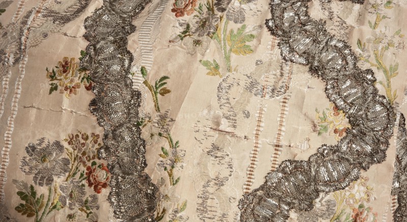 Robe à la française robe à paniers à coudes, two-piece, pink grosgrain with swirling silver lamellar brocade, silver lace ribbons and bouquets of flowers; c. 1764. Worn on an oval corselet. a Outer dress manteau open at the front with pleated train at the back. Bodice stiffened with 22 long bonings, laced at front and back under the pleats. Elbow-length curved sleeves finished with double flounces trimmed with silver lace. Constructed from eight panels; quadruple pleats at the back preserve full fabric width, with cuts and fittings only at the front. Silver lace appliqué on the edges echoes the wavy brocade pattern. b Apron of same fabric, hemmed with a wide ruffle and matching silver lace appliqués repeating the wavy brocade motif. Materials/measurements: fabric height 37 cm x width 27 cm; fabric width 55.5 cm. Garment length 167 cm; width at bottom of manteau 430 cm. Origin: France fabric; place of use: Poland., Tags: lace ornaments; floral bouquets; ribbon ornament; women's 18th-cen...