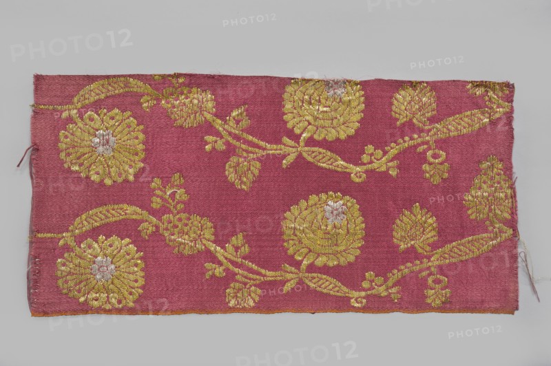 Fabric fragment, fragments of silk patterned semi-silk fabric with a red background in a satin weave and brocaded decoration using two types of braided metal thread with yellow and white cores. Vertically arranged pattern of a waving, fanciful flowered and leafy branch with small leaves and large oval heads of stylized pompon flowers alternating in horizontal rows. Dyes: indigo, brazilwood. Semi-silk satin, brocaded. Weaving edge: 0.3 cm wide, orange cotton warp and cherry cotton weft in plain weave. Construction: satin warp weave; thicker red cotton wefts and red silk warp giving stiffness. Pattern introduced with brocade wefts using two types of loosely braided threads – a gold plate on a yellow cotton core and a silver plate on a white cotton core. Continuous longitudinal pattern on a red background of symmetrical trails of bending gold twigs with leaves, fruit, and two types of flower heads with silver centers. Pattern repeat width 10.5 cm. Selvedge width 0.3–0.4 cm. I-thread: yell...