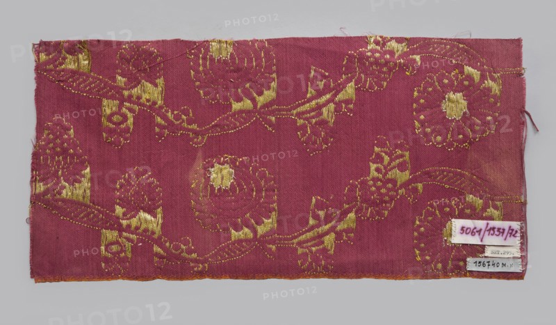 Fabric fragment, fragments of silk patterned fabric with plant motifs, indigo and Brazilwood dyes. Semi-silk with a red satin-weave background and brocaded decoration using two types of braided metal thread with yellow and white cores. The vertically arranged pattern shows waving, fanciful flowered and leafy branches covered with small leaves and large oval stylized flower heads alternating left and right in horizontal rows referred to as pomponblüten. Floated and brocaded with double metal thread gold plate on yellow core and silver plate on white core. Continuous pattern along the length on a red background—symmetrical trails of regularly bending gold twigs with leaves, fruit, and two types of flower heads with silver centers. Construction: satin-warp weave; thicker red cotton wefts and red silk warps give stiffness. Weaving edge 0.3–0.4 cm wide with an orange cotton warp and cherry cotton weft in plain weave. Pattern repeat c. 10.5 cm. Tapestry-like brocaded satin, likely 17th centu...