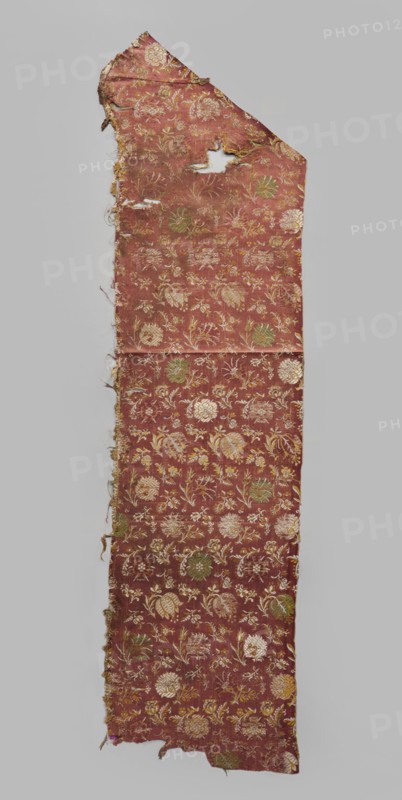 Fabric fragment, 17th century, Italy, warp-woven satin background with pale red silk warp and two alternating wefts white and yellow creating the pattern using the lisère technique; brooching wefts in green and beige; high fabric tightness. SATIN WITH A TWO-COLOR WEFT, LISÉRE. Construction: floating runs in diagonal, fairly wide bands. Pattern report length 29 x width 17.5 cm. Floral motifs in three types arranged at close intervals: two kinds of carnations rows facing left or right, tulips with drooping calyxes, and roses with spread petals, with smaller flower projections scattered between them facing in different directions. Colors of flowers and elements vary — white and yellow stems and leaves, green and beige calyxes. Fragments of silk patterned fabric, height 112 cm, width 28.5 cm., A long, narrow piece of worn fabric with an irregular top edge and frayed sides, patterned with a repeating floral motif; the fabric's base color is a muted reddish-brown with floral elements in shad...
