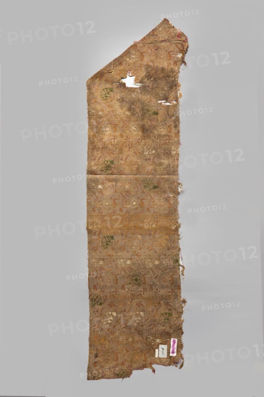 Fabric fragment. 17th century, Italy . Warp-woven satin background of pale red silk warp with two alternating wefts white and yellow forming the pattern by the lisère technique—floating runs in diagonal, fairly wide bands. Brooching wefts: green and beige. High fabric tightness. Pattern report 29 x 17.5 cm. Floral motifs in three types, arranged closely: two kinds of carnations rows facing left or right, tulips with drooping calyxes, and roses with spread petals, with smaller flower projections scattered between them, facing different directions. Flower colors and elements vary—white and yellow stems and leaves, green and beige calyxes. Materials: silk; satin with a two-color weft. Dimensions: height 112 cm, width 28.5 cm., A long, narrow, irregularly shaped piece of fabric with frayed and torn edges, shown against a plain light gray background; the fabric has a faded golden-brown base color with a repeating floral and foliate pattern woven in lighter gold and muted green, and accents ...