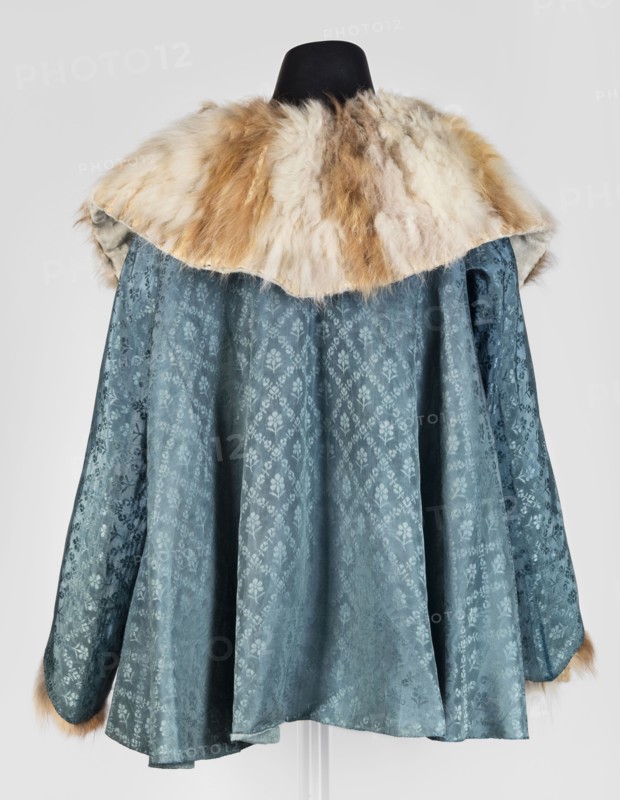 Jubka, long women's caftan jubka, hip-length, narrow in the shoulders, flared at the bottom; late 18th century, Poland. Large turn-down collar entirely trimmed with fur two types of fur decoratively connected in narrow bands. Long set-in sleeves, narrow at the top, flared at the bottom, lined with white sheepskin with strongly twisted fleece. Geometric ornaments; women's clothing, fashion. Dimensions: length 63 cm, circumference 292 cm at the bottom., A garment displayed on a black mannequin with a wide fur collar in shades of cream, beige, and light brown, draped over a flowing fabric body in a muted teal-blue with a subtle floral damask pattern, the sleeves and hem showing the same teal-blue fabric and small touches of the light brown fur at the cuffs, all against a light gray background.