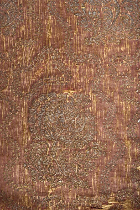 Fabric fragment, silk brocade grosgrain, 1st half of the 17th century, Italy. On a red background a gold floral pattern of projections on a curved leafy stem with a large stylized tulip calyx; motifs arranged in rows shifted by half a repeat, alternating lean left and right, with a wavy twig with small leaves between them. Construction: yellow silk weft, red silk warp in grosgrain weave; brocaded thread of gold plate braiding on a yellow silk base. Pattern repeat: 33 cm x 17 cm. Border width: 1 cm. Colors: yellow-brown and gold threads. Dimensions: 39.5 cm high x 43 cm wide., The image shows a textured fabric with an intricate woven pattern featuring floral and swirling motifs. The dominant colors visible are reddish-brown, gold, and muted green, with highlights of pale gold or beige creating a slightly metallic sheen. The surface displays a mix of vertical striations and raised embroidered-like areas where the pattern is more pronounced, and there are a few small worn spots where the ...