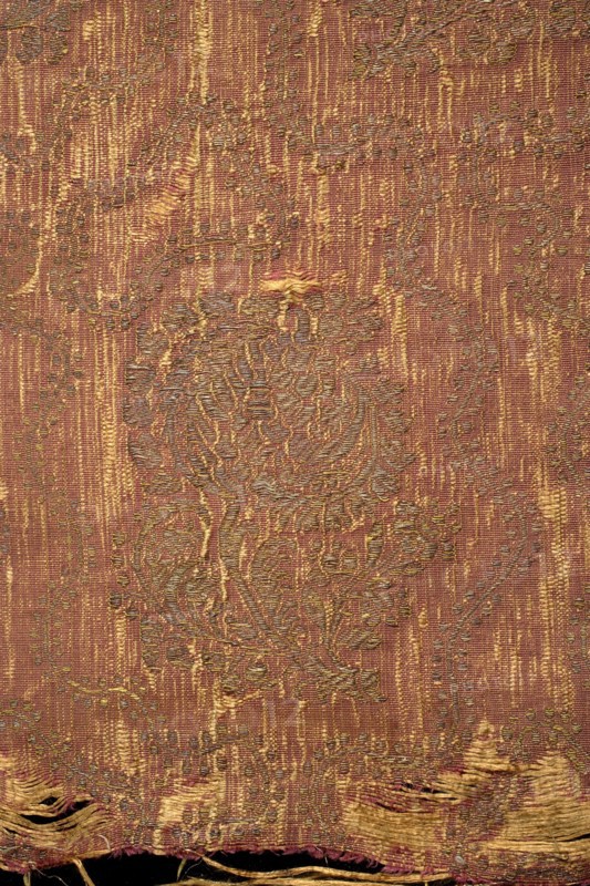 Fabric fragment, silk brocade brocaded grosgrain, 1st half of the 17th century, Italy. Red ground with a gold floral pattern of projections on curved leafy stems, featuring a large stylized tulip calyx. Motifs arranged in offset rows shifted by half a repeat, alternating lean left and right; between them a wavy twig with small leaves. Construction: yellow silk weft, red silk warp in a grosgrain weave; brocaded thread of gold plate braid on a yellow silk ground. Pattern repeat: 33 cm × 17 cm. Border width: 1 cm. Colors: yellow-brown, gold. Overall dimensions: height 39.5 cm, width 43 cm., A close-up of a woven textile showing a worn patterned surface with a vertical striated background and an overlaid ornate floral motif; the fabric displays frayed edges and threadbare areas near the bottom, with visible loose threads and small gaps. The colors visible are shades of reddish-brown, gold, and muted olive, with lighter golden highlights in the raised embroidery and darker brownish-red thre...