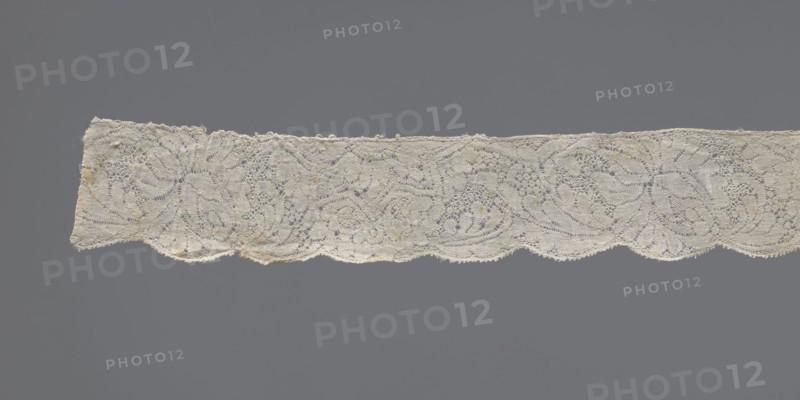 Lace fragment, early 18th century, Binche. Plant ornaments with rosette and rose motifs; twig motif with large leaves and flowers. Upper straight edge has half stars with rosettes inside; lower edge is gently serrated. Background varied. Lace fabric; width 3 cm, length 130.5 cm., A horizontal strip of delicate lace with a scalloped lower edge stretches across a gray background; the lace is off-white with small areas of light tan discoloration, featuring an intricate openwork pattern of swirling floral and leaf-like motifs, fine looping threads, and tiny holes, with slightly irregular edges and a few loose fibers visible.