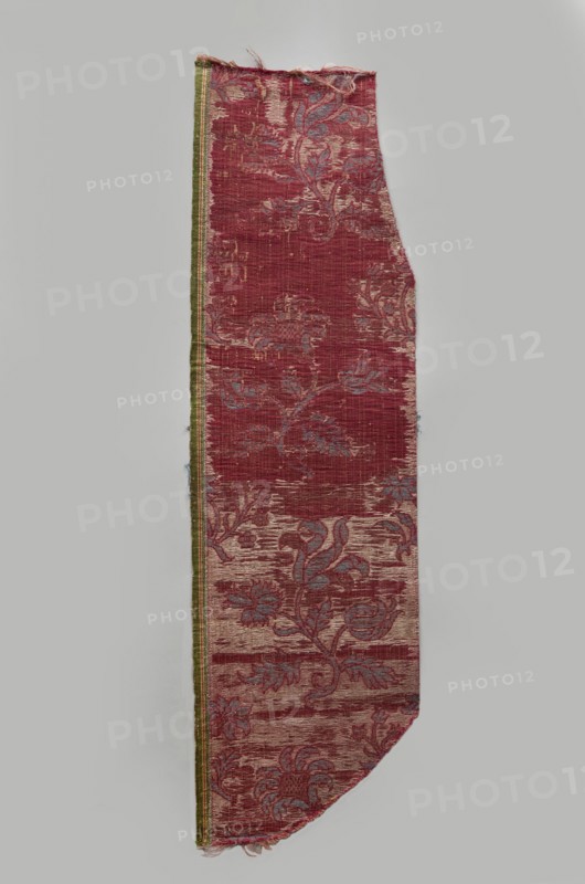 Fragment of fabric, silks, brocade, floral and plant patterns, double-warp brocade. On a white and silver background, blue motifs with a cherry outline. On a white and cherry background, densely placed motifs of blue flower bushes with a cherry outline — sunflower-like flowers on leafy bent stems, alternating in rows shifted by half the motif width, and S-shaped flower branches with a tulip and a carnation. Fragmentary elements suggest additional smaller motifs between the two main ones. Construction: background in plain warp weave - basic linen thread, bold cherry-red silk warp; additional pink silk warp binds in a twill weave the patterning threads: silk blue pattern and white background covering, plus bundles of two silver lamellas running on the surface across the fabric width. Pattern repeat: length 45 cm. Weaving edge: 1.3 cm wide in green with yellow, pink and white stripes. Date: 16th/17th century, Italy. Dimensions: height 77 cm, width 21.5 cm., A long, narrow textile scrap wi...