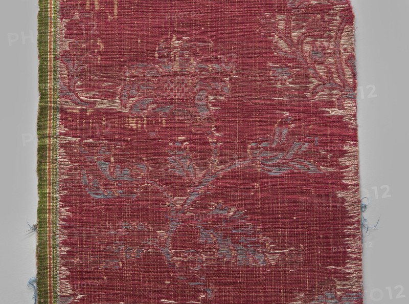 Fragment of fabric, silks, patterned brocade with floral and plant motifs, double-warp brocade, 16th/17th century, Italy. On a white and silver background, blue motifs with a cherry outline. On a white and cherry background, densely placed motifs of blue flower bushes with a cherry outline: sunflower-like flowers on a leafy bent stem, alternating in rows shifted by half the motif width, and S-shaped flower branches with a tulip and a carnation; fragmentary elements suggest additional smaller motifs between the two main ones. Construction: background in a plain warp weave of basic linen thread with bold cherry-red silk warp; additional pink silk warp binds in a twill weave the pattern threads: silk blue pattern and white background covering, plus bundles of two silver lamellas running on the surface across the full width. Pattern repeat length: 45 cm. Weaving edge: 1.3 cm wide in green with yellow, pink and white stripes. Dimensions: height 77 cm, width 21.5 cm., A worn woven textile fr...