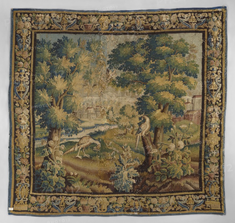 Tapestry-verdure, In the foreground, three flowering shrubs; behind them two birds, one perched on a tree trunk and one fluttering between the branches; two large trees. In the background, a landscape with architecture and a river. Border with motifs of flower bouquets in vases. Floral ornaments, flowers, leaves, trees, birds, landscapes, borders. Colors: green, sand, sky blue, dark blue. 17th century, Flanders. Woven fabric; height 272 cm, width 278 cm., A woven tapestry showing a landscape scene with a group of large leafy trees and a few smaller plants in the foreground, rendered in shades of green, gold, brown, blue, and cream. Near the center foreground, a bird perches on a tree stump while another bird feeds on the ground among grassy mounds and foliage in green and brown tones. In the midground a flowing waterway winds past grassy banks toward a distant area with indistinct built structures partially obscured by trees. The tapestry has a decorative border featuring floral and fo...