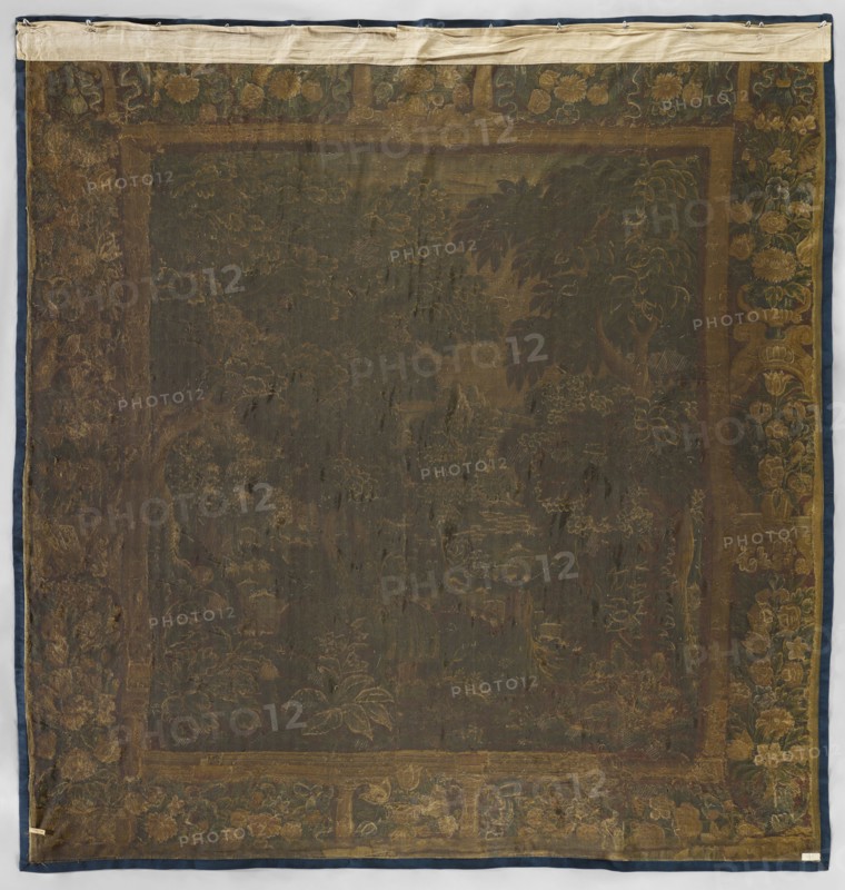 Tapestry-verdure, In the foreground are large trees and blooming shrubs and flowers; further on a river; beyond the river are rural buildings. Border: side panels with vases of flowers; horizontal panels with flower garlands; center with vases of flowers. Colors: sand, brown, blue, green, and ochre. 17th century, Netherlands. Woven decorative fabric tapestry, height 319 cm, width 308 cm., A large square textile with a dark central field surrounded by a patterned border; the fabric shows a faded floral and foliage design with leaves and flower shapes rendered in muted gold and brown tones against a deep greenish-brown background, and a surrounding band featuring floral motifs in shades of gold, olive, and dark green with touches of brown; the outermost edge is trimmed in a dark blue fabric and the top edge has a lighter beige strip attached; the surface appears worn and uneven in texture with areas of lighter and darker discoloration and visible weave.