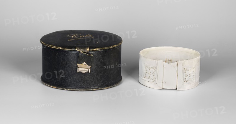 Women's collar, made of thin white linen, stiffened, decorated with openwork embroidery; edges on the front decorated with rosettes in the form of a square. Collar, white embroidery, clothing accessory, 19th/20th century 19th–20th c., Poland; height 5.5 cm, width 40 cm., A dark round case with worn edges and a small metal clasp sits next to a light-colored cylindrical fabric band featuring decorative cutwork panels and a folded closure; the scene shows shades of black, off-white, cream, gray, and touches of metallic gold on the case against a neutral gray background.