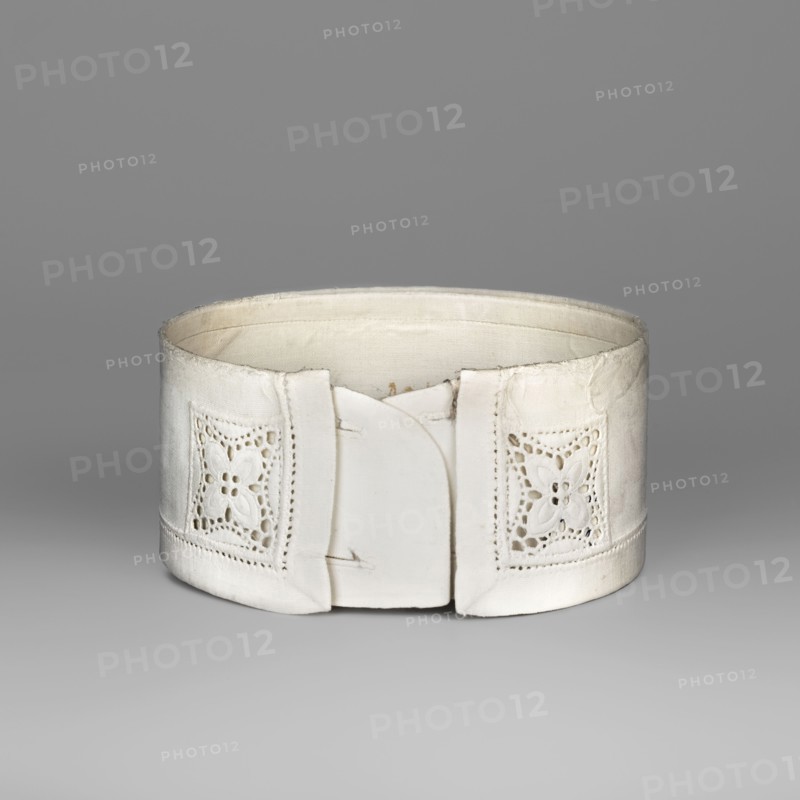 Women's collar, made of thin white linen, stiffened, decorated with openwork embroidery. Edges on the front are decorated with rosettes in the form of a square. White embroidery. 19th/20th century, Poland. Clothing accessory; height 5.5 cm, width 40 cm. Function: women's fashion accessory collar., A white cylindrical fabric object with a cutwork floral motif on each side and a plain central front panel, shown against a smooth gray background; the fabric appears slightly worn with subtle shadows and highlights in shades of white, off-white, cream, and gray.