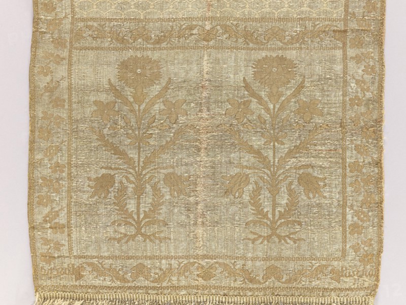 Kontusz sash by Paschalis Jakubowicz Manufactory Warsaw; belt manufacturer; 1788–1794. Bipartite heads with floral bouquets. Still with carp scale ornament. Border with floral and plant twig. Sewn-on fringes. Ornament - Heads: two motifs of rooted flower bushes with a carnation flower at the top. - Center: carp scale motifs. - Edges: vertical motifs of flower branches forming a twig; horizontal twig motif with palmettes. - Borders: checkerboard. Coloring - Right: silver background, light brown ornament of heads and edges, cream carp scale design. - Left: reverse of right. Signed at the bottom in the heads: Paschalis. Date: 18th century 1788–1790. Place of origin: Lipków, Warsaw . Type: national costume accessory kontusz sash. Dimensions: length 372 cm, width 29 cm., A rectangular woven textile with a repeating floral pattern in shades of gold and beige, showing a symmetrical design of stylized flowers with tall stems and leaves against a slightly lighter background, framed by a decorat...