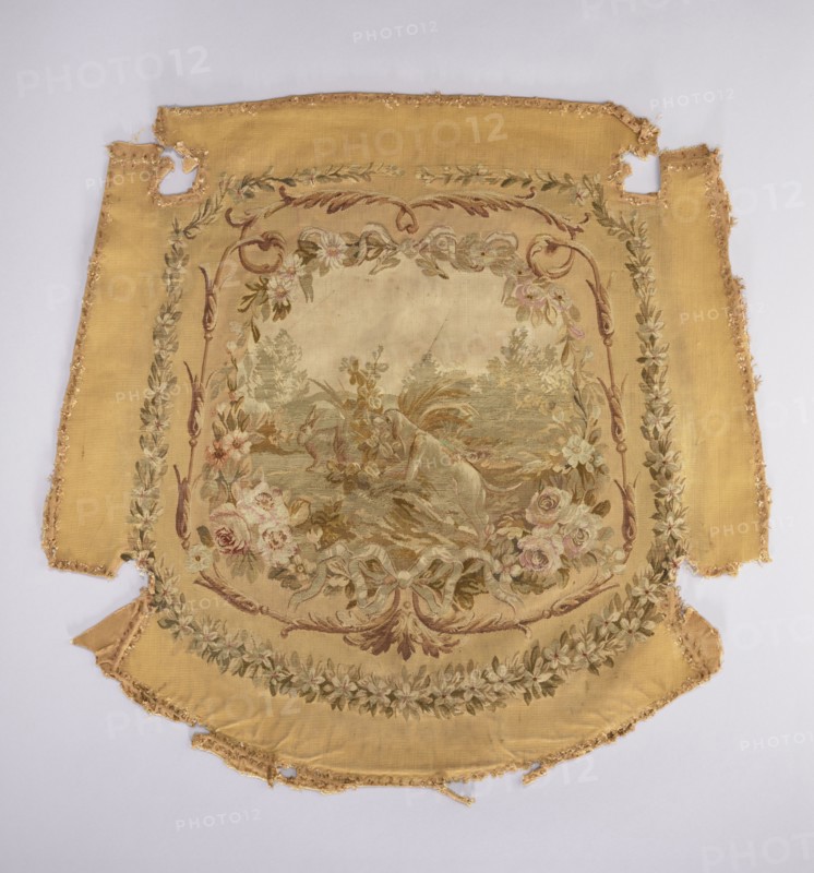 Tapestry hangings, varietals, message provenance, flowers, floral ornaments, fabrics, tapestries, interior furnishings, decorative fabrics, weaving, dogs, female figures, figures, furniture upholstery, genre scenes against a landscape background. Seat with a depiction of a dog sitting on grass, stage surrounded by a floral and acanthus border. Predominant colors: beige background, green, pink, and brown. The backrest depicts a young woman in a theatrical costume against a garden background. The scene is framed by a floral and acanthus border in the same tone as the seat. Armrests with a garland of blooming jasmines. 18th century, France. Fabrics. Height 75 cm, width 78 cm., A large worn textile panel with a central painted scene framed by ornate floral and ribbon motifs, showing a muted palette of gold, beige, cream, green, brown, pale pink, and soft red tones; the outer field is a warm golden yellow with frayed and torn edges and a few missing sections, while the central area is light...