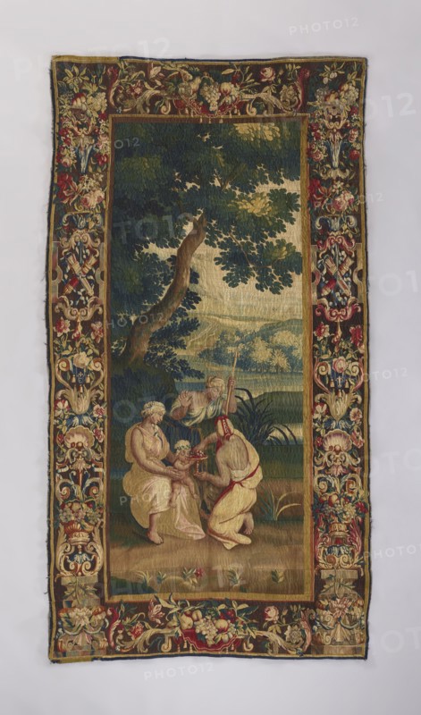 Figural tapestry, flowers, floral ornaments, Dionysus mythology, mythological scenes, mythological figures, figures of women with children, weaving, interior furnishings, decorative fabrics, interior decorations, Bacchic scenes, tapestries, nymphs. In the center of the landscape, little Dionysus kneels at the feet of one of the nymphs; another pours wine into his goblet, a third stands nearby with a spear. The depiction is surrounded by a border with motifs of flowers, acanthus leaves, shells, and ogee. Colors: brown, green, red, blue, ochre, and sand. 18th century, Brussels; fabrics; height 256 cm, width 136 cm., A rectangular woven tapestry with a decorative floral border in red, gold, green, blue, beige, and cream surrounds a central pastoral scene; within the central panel, a large tree with dark green and olive leaves and a brown trunk dominates the upper area against a background of green and blue distant hills and a pale beige sky, while in the foreground a group of figures wear...