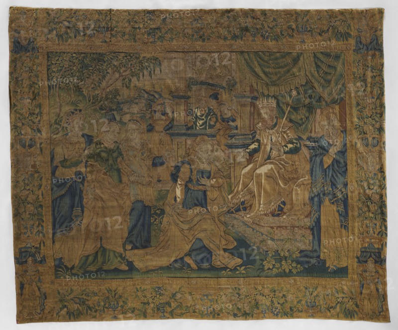 Figural tapestry; late 16th century, Flanders. On the right, a king wearing a crown and holding a scepter sits on a throne under a canopy, next to courtiers. A young woman kneels before the king, and behind her are three ladies-in-waiting. In the center background, terraced buildings with other courtiers; a distant landscape beyond. Surrounded by a border with floral and leaf motifs; in the corners, under canopies, female figures. Materials/technique: woven tapestry; fabrics. Colors: green, sand, blue, yellow, and ochre. Dimensions: height 296 cm, width 355 cm., A woven tapestry showing a group of robed figures gathered around a seated crowned figure on a throne, all depicted in muted golds, deep blues, and faded greens; the central seated figure wears layered garments in gold and dark green and is positioned on a raised platform with drapery behind in green tones, while surrounding figures wear flowing robes in gold, blue, and green and display varied poses and gestures; the scene is ...