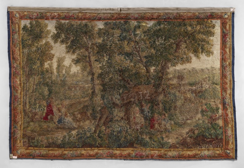 Figural tapestry, Chodecz Kuyavian-Pomeranian Voivodeship; flowers, floral ornaments, dogs, children, women; genre scenes against a landscape background; tapestries, interior furnishings, pastoral scenes, decorative fabrics; In the foreground a group of children playing against a background of tall trees. On the right a girl with a shepherd's staff sits on the grass and a young man next to them a dog. In the background on the right and left there is architecture. Narrow border with a motif of a simple frame entwined with flowers; cartouches in the corners. Colors: green, red, blue, yellow, brown, ochre, and cream. 2nd half 19th century, Germany; fabrics; height 265 cm, width 395 cm, A large rectangular woven tapestry hung flat against a light background, showing a wooded landscape scene with a central group of tall trees with green and olive foliage and brown trunks, surrounded by grassy and shrubby undergrowth in shades of brown, tan, green, and muted blue. Along the edges, a decorati...