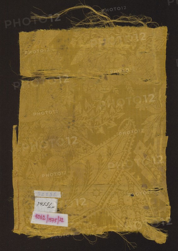 Fabric fragment, patterned silk damask, early 18th century, France. Yellow semi-silk damask with floral-fantasy motifs; background is warp-covered satin with a plain-weave ornament. Decorated with stylized large leaves with frayed edges, small heart-shaped leaves, small leaves and flowers, blades of grass, small fruit, details evoking Far Eastern architecture, and openwork lace-like elements in a bizarre style. Color: Jaune d'or Newton. Pattern scale larger than preserved fragment. Dimensions: height 34 cm, width 23.5 cm., A rectangular piece of worn fabric with frayed edges and loose threads displayed against a dark background, the fabric showing an intricate woven floral and geometric pattern visible through texture and subtle sheen, with areas of thinning and small holes and a horizontal tear near the top; the dominant color of the fabric is golden yellow with variations in tone, and there are a few small labels attached near the lower left corner that contain handwriting and printe...