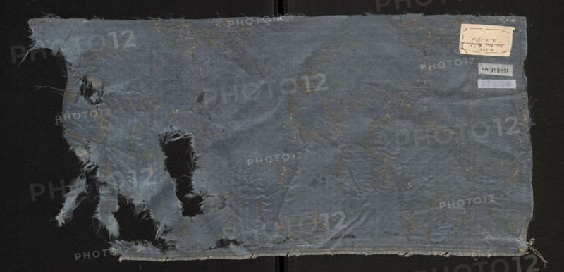 Fabric fragment, fragments of silk fabrics, patterned fabrics. Report composition based on two intersecting diagonal arrangements: one formed by ribbons winding wavy-like from tiny, fancy leaves brocaded with silver thread; the other running in the opposite direction with forms resembling sheaves brocaded with gold thread. Satin background enriched with a miniature rhomboidal net pattern in liseré. Blue Gros de Tours background with elements of liséré. Brooch motifs: silver lamellar and gold-braided thread with a loose braid showing the silk core in natural color. Report size larger than the preserved fragment. Motifs in the bizarre fabric type. Multicolored border: green, pink, and yellow threads. Late 17th century, France. Height 56 cm, width 27 cm., A worn rectangular piece of fabric laid flat with frayed edges and several irregular holes, showing loose threads and torn strips along one side; the fabric is primarily a muted blue with patches of darker blue where folds and wear appea...