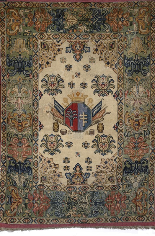 Rug with the Potocki and Mniszech coats of arms. A light central field surrounded by a wide three-band border; the widest band has a palmette motif. In the central field is an octagonal coat-of-arms cartouche: on the right the Pilawa coat of arms on a blue background, on the left the Konczyce coat of arms on a red background; above the cartouche a noble crown; behind the cartouche two crossed hetman's maces and also swords, pikes, banners, and cannon barrels. Below the cartouche are symmetrically arranged pairs of barrels and cauldrons, and two stacks of cannonballs. Possibly 1st half of the 18th century, Brody, Poland. Fabrics; height 209 cm, width 142 cm., A rectangular woven textile with a central cream field surrounded by a wide patterned border, featuring a central shield motif in red and blue topped by a gold crown and flanked by stylized banners in blue and tan; the cream field contains several floral rosette motifs in blue, tan, red, and green and pairs of symmetrical stylized ...