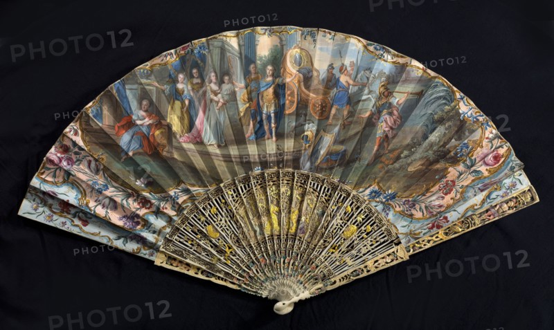 Fan, double-sided fan, paper painted with gouache and gold dust. On the obverse, a scene of two knights dressed in Roman costume welcomed by trumpeters; women in flowing robes greet them, one carrying a child. Composition surrounded by a margin of leaves and flowers; decorated in light pastel colors and gilded ornament. On the reverse, children playing in a meadow among trees and flowers. Feathers cut from ivory, openwork, painted with oil and inlaid with gilded tin foil. Central cartouche with a figural representation against palace and garden architecture. 2nd half of the 18th century, France. Height 40.4 cm., A decorative folding fan displayed open against a dark background shows a painted scene across the leaf with a group of figures in period clothing and classical architectural and landscape elements, framed by floral ornamental borders; colors visible include blue, pink, yellow, gold, green, red, brown, cream, and black. The fan sticks and guards feature intricate openwork carvi...