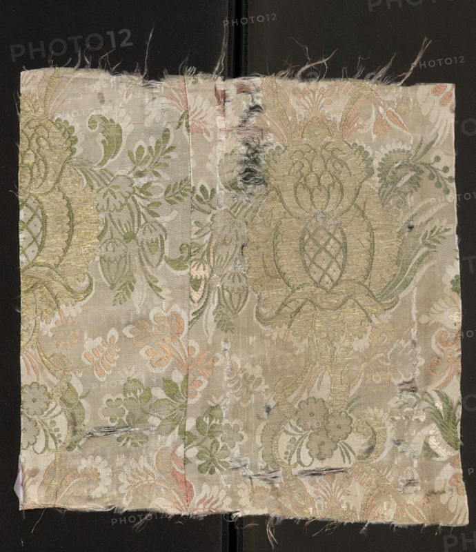 Fabric fragment. Cream silk damask fabric, embroidered and brocaded with colorful threads. Promoting threads: green, light and dark salmon. Brooching thread: gold. Pale cream satin background with weft weave coverings, embroidery, and brocade. Fantastical floral decoration: small twigs and leaves of the damask sub-report complement larger motifs brocaded with gold threads. Stylized pomegranates pineapples placed on a winding stem, surrounded by sprigs of smaller flowers. Fabric sewn from two pieces. Composition of the report motifs based on mirror symmetry. Weave: satin weave with warp coverage; pattern creating a sub-report in a plain weave; colorful pattern promoted in zones; promoting threads tied in a twill weave introduced two at a time in the interlacing. Date: 1710–1715. Place of origin: France. Materials: silk, metal thread. Dimensions: height 33 cm, width 33.5 cm., A rectangular piece of fabric with frayed edges resting on a dark background shows an ornate floral and foliate w...