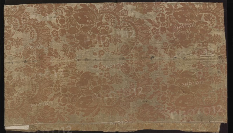 Fabric fragment. Fragment sewn from two salmon-white damask-type fabrics promoted stripes, differing slightly in pattern and ply width. 1. Damask effect achieved by different colors of warps and wefts. Pattern in mirror symmetry. Tiny blossoms spill out from large, blooming peony-type flowers. White taffeta background, salmon pattern — gros de Tours. Report: height 29 x width 49.5 cm; edge width 0.5 cm. 2. Damask effect achieved by different colors of warps and wefts. Pattern in mirror symmetry. Large fruit and blooming flowers, lace motifs resembling combs of French needlepoint lace. White taffeta background, salmon pattern — gros de Tours. Report: height 38.5 x width 50.5 cm; edge width 0.4 cm. Dated 1735–1745, France. Overall fabrics: height 98 cm, width 97 cm., A rectangular piece of fabric laid flat, showing a repeating floral and paisley-like pattern in a muted reddish-brown against a faded beige-gold background; the cloth has visible horizontal fold lines and some darker smudges...