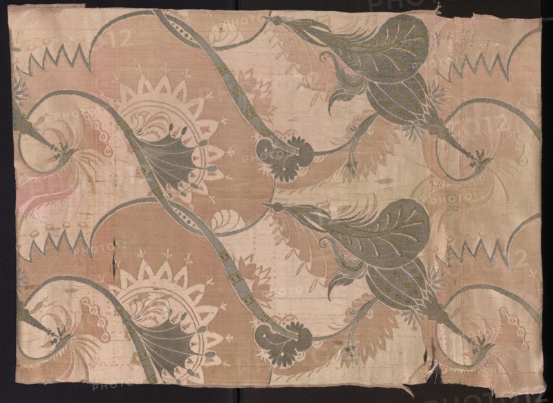 Fragment of pink silk brooch fabric with a fanciful and floral pattern in the Bizarr type. Early 18th century, France. Pattern: arrangement of intertwining tendrils with fanciful forms—some half-vegetal, half-animal fantastic winged dragons, others topped with sharp edges like poppy-head shafts. Construction: satin with warp cover and simple weave background; other motifs brocaded with metal threads. Colors: pink ground; brocades in silver, green, pale pink; silver metal thread with a loose braid. Width of fabric: 54.5 cm. Report dimensions: height 60 x width 27 cm; overall height 106 cm, width 54 cm., Classification: patterned silk fabric, fragment., A rectangular piece of patterned fabric with a faded background in shades of pale pink and beige, featuring a repeating ornamental design of flowing curved vines and large stylized leaves in muted gray-green with small olive accents. The fabric shows a few scalloped circular motifs in off-white and pale tan, and several areas of paler bei...