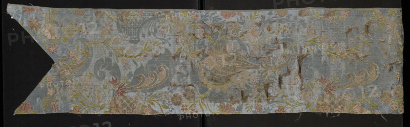 Fabric fragment, origin unknown, early 18th century, France. Silk lisère warp satin fragment cut in the form of a chasuble column with a triangular neckline; warp-woven lisère satin with prominent brocaded pattern. Right side: damask subrapport of lisère satin background and plain weaves decoration; left side: plain weaves. Decorated with whimsical plant motifs: tiny flowers on slender celadon-green branches and fanciful forms resembling onion-shaped flower buds growing from ornamentally treated horn-shaped bases; lace-like patterning. Colors: blue background with colorful brocade and embroidered threads. Brocade threads: gold, white, pale pink, blue, burgundy; embroidered threads: orange, pink, blue. Pattern created by threads tied in a plain/simple weave. Material: silk, lisère satin, embellished and brocaded. Dimensions: height 106 cm, width 23 cm; repeat report length 44 cm., A long horizontal piece of fabric with a pale blue background featuring an ornate floral and scroll pattern...