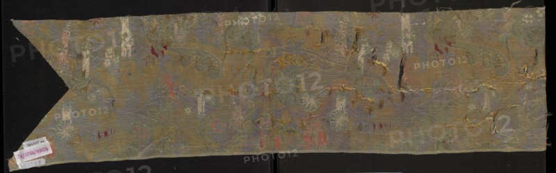 Fabric fragment, origin unknown, early 18th century, France. Fragment cut in the form of a chasuble column with a triangular neckline. Warp-woven liséré satin with a prominent brocaded pattern: blue warp-satin ground with colorful brocade and embroidered threads. Pattern composed of whimsical plant motifs — tiny flowers on slender celadon-green branches and fanciful forms resembling onion-shaped flower buds growing from an ornamentally treated horn-shaped base — and lace-like patterns. Brocade threads: gold, white, pale pink, blue, burgundy; embroidered threads: orange, pink, blue. Right side shows a damask subrapport of liséré satin background and plain weaves decoration; left side has plain weaves. Threads tied in a plain/simple weave. Materials: silk, liséré satin, embellished and brocaded. Report length: 44 cm. Dimensions: height 106 cm, width 23 cm., A long, narrow piece of woven fabric displayed against a dark background, showing a faded, ornate floral and foliate pattern with fl...