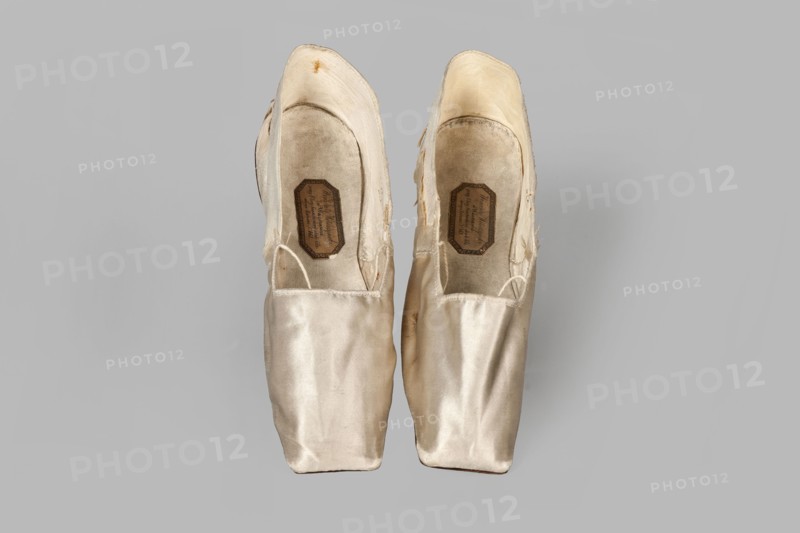 Women's slippers by Wincenty Witoszynski, ca. 1840–1850. Shallow, flat women's slippers made of white silk. Symmetrical thin leather soles with straight-cut toes. Lining: front of white canvas; back of thin white leather. Inside an octagonal company label: Wincenty Witoszynski / in Warsaw / at Senatorska Street next to the Main Town Hall No. 463. Upper part machine-sewn. Materials: silk, leather, canvas. Dimensions: height 5.3 cm; width 5.3 cm; length 23.2 cm., A pair of worn ballet pointe shoes seen from above, placed side by side on a neutral grey background; the shoes are a pale ivory to cream color with satin fabric showing creases and slight discoloration, the interiors appearing a bit darker with beige and tan tones, and there are visible areas where the fabric is frayed and stitched, along with a few straps of matching color looped inside.