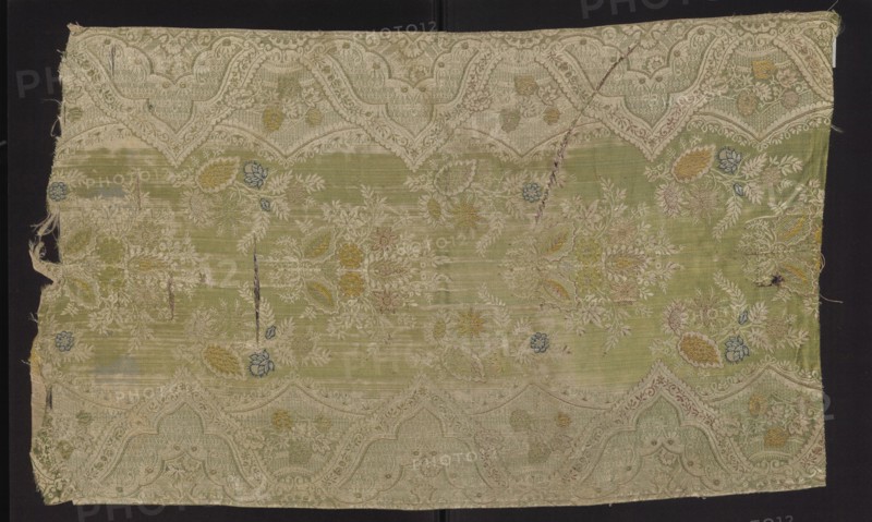 Fabric fragment, broached silk damask, early 1st quarter of the 18th century, France. Warp-covered satin ground with green warp. Broaching wefts in light and dark pink, pale and dark blue, celadon, yellow, and salmon; feature threads dark and light pink and white, captured in a weft weave. Unilateral two-color damask effect, with mirror symmetrical lace pattern: floral sprigs along a central axis and serrated lace border with floral bouquets. Stylized yellow and blue-green flowers on a light green background. Preserved selvedge, pile width, and pattern repeat. Pattern width 50 cm; repeat height 48 cm, repeat width 48 cm; edge width 0.6 cm. Fragment dimensions: height 79 cm, width 48 cm., A rectangular textile fragment with a faded pale green ground and an ornate woven border in light beige forming scalloped arches along the top and bottom edges, decorated overall with floral and foliate motifs; the surface shows several embroidered or woven flower clusters and leaves in muted yellow, p...
