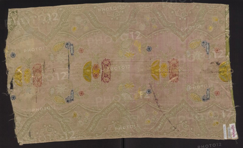 Fabric fragment, silk brocaded and broached damask, 1st quarter of the 18th century, France. Warp-covered satin ground with green warp. Broaching wefts in light and dark pink, pale and dark blue, celadon, yellow, and salmon; feature threads dark and light pink and white, captured in a weft weave. Unilateral two-color damask effect, broached with colored wefts. Lace-style motif arranged in mirror symmetrical composition: floral sprigs along a central axis and a serrated lace border with floral bouquets. Pattern width 50 cm; repeat height 48 cm, width 48 cm; edge width 0.6 cm. Fragment dimensions: height 79 cm, width 48 cm., A rectangular piece of woven fabric laid flat with a faded pink background and a subtle repeating scalloped pattern in a lighter beige tone, showing areas of wear and small holes. Scattered across the surface are embroidered or woven floral and abstract motifs in several colors including yellow, blue, red, green, and gold, with some motifs appearing as semi-circular ...