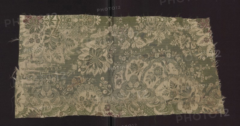 Fabric fragment, early 18th century, France. Silk fabric with preserved patterned and lace-like designs, featuring plant and floral motifs. Colors: green background with sand-beige and cherry weft. Decoration: large repeat with symmetrical, mirror-layout motif; repeat size 48.5 x 49 cm. Dimensions: height 52 cm, width 24.5 cm., A rectangular piece of woven fabric with frayed edges laid on a dark background, showing an overall floral and foliate pattern with large stylized blossoms, leaves, and decorative scalloped motifs; the fabric displays a mottled appearance with areas of wear and small holes, and threads visible along the cut edges. The visible colors include green, beige, off-white, and small touches of muted red and brown.