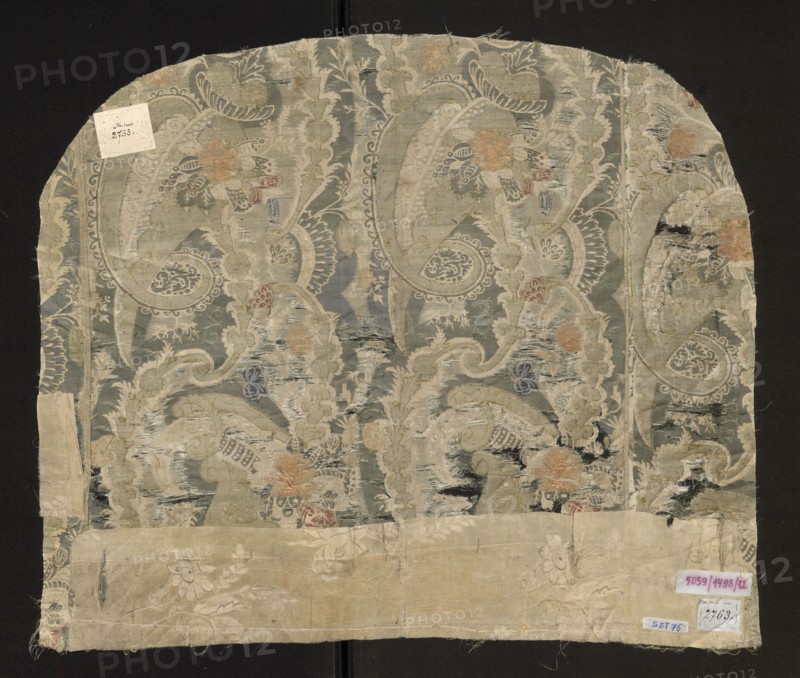 Fabric fragment, fabrics, fragments of a larger textile object, fragments of silk fabrics, patterned fabrics. Flaunted and brocaded silk. Damask subreport - background in a satin weave, pattern in a grosgrain weave, green flaunting thread tied in a twill weave. The white, green, and gold pattern provides a backdrop for colorful floral and fanciful motifs. The asymmetrical fabric report is created by larger forms resembling cornucopias, which intertwine turbinate, lace, and plant motifs. Bizarre decoration. Flaunting thread - green. Padding threads: salmon, pink, blue, gold braided thread. Beginning of the 18th century, France, fabrics. Dimensions: height 51 cm; width 61 cm; width of the brim 43 cm; width of the edge 0.3 cm; width of the report 23 cm., A worn rectangular textile fragment with an arched top displayed against a dark background, showing an ornate repeating paisley and floral design in muted colors including grayish blue, beige, pale green, soft orange, faded red, and hints...