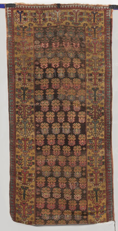 Carpet, gift; 18th century, Caucasus. Main field with floral branches arranged in alternating rows. Two-band border: wide inner one with flowering tree motifs and narrow outer one with a tendril. Colors: brown, yellow, and red backgrounds. Motifs are highly geometric. Materials: fabric; height 219 cm, width 111 cm., A rectangular woven textile with an overall pattern of repeating floral-like motifs in rows on a dark brown field, surrounded by a wide mustard yellow border that contains geometric, cross-like designs; the textile shows colors including dark brown, mustard yellow, red, pink, blue, teal, green, and small areas of cream, with visible wear and slight fading in places, fringed edges at the top and bottom, and narrow side bands with alternating small geometric elements.