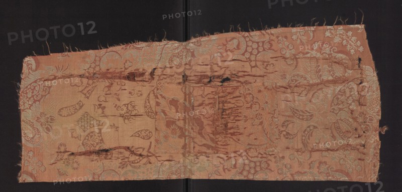 Fabric fragment, fragments of a larger textile object, patterned silk fabrics. Pale red pink lampas with a white patterned weft — a unilateral two-tone damask. Background: light red/pink warp satin; overlays in plain and liseré weaves with a white weft tied in a weft weft-faced weave; reverse side in plain weave. Repeat rapport composed of small flowers, fanciful leaves, and miniature lace motifs forming an ornament on the leaves; motifs typical of bizarre fabrics inspired by lace. Another fragment likely part of the same fabric shows a pale red stripe with a white pattern weft. Subreport: plain weaves pink warp/pink weft and white weft weaves. Colors: pink background with white decorative patterning. Hem width: 0.4 cm. Rapport: length 64.5 cm; width 24.5 cm. Overall fabric height 73 cm, width 31 cm. Early 18th century, France., A rectangular piece of worn fabric with a faded floral and paisley woven pattern rests against a dark background; the textile shows frayed edges and several lo...