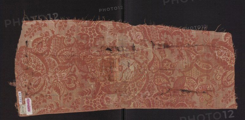 Fabric fragment, fragments of a larger textile object, patterned silk fabrics, pale red pink lampas with a white patterned weft. A unilateral two-tone damask: light red/pink warp satin background, overlays in plain and liseré weaves with a white weft tied in a weft weft-faced weave; reverse in plain weave. Repeat composed of small flowers, fanciful leaves, and miniature lace motifs forming an ornament on the leaves; motifs typical of bizarre fabrics inspired by lace. Fragment no. SZT 79 likely part of the same fabric. Weave structure details: background in warp satin pink warp; subreport areas in plain weave pink warp, pink weft; white weft areas in weft weave; reverse plain weave. Colors: pink background with white decorative pattern. Hem width: 0.4 cm. Repeat report dimensions: length 64.5 cm; width 24.5 cm. Overall dimensions: height 73 cm; width 31 cm. Date: early 18th century, France., A rectangular piece of woven fabric with frayed edges lies flat against a dark background, showi...