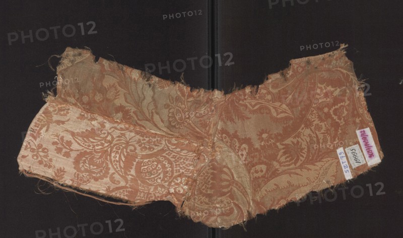 Fabric fragment, Patterned silk in a two-color damask type with a decoration of stylized leaves. Stripe weave structure, pale red accented with a white patterning weft. A type of unilateral two-color damask. Background — light red warp satin, overlaid with plain and liseré weaves, accented with a white weft tied in a weft weave; reverse side in a plain weave. The repeat is composed of small flowers, fanciful leaves, and miniature lace motifs forming an ornament on the leaves. The repeat dimensions are larger than the preserved fragment. Motifs in the bizarre fabric type inspired by lace. Fabric fragment no. SZT 78 is probably part of the same fabric. Hem width: 0.5 cm. Early 18th century, France. Height 53 cm, width 19 cm. Materials: silk. Classification: fabrics, fragments of patterned silk., A worn textile fragment with a decorative floral and paisley pattern sits against a dark background; the fabric shows frayed edges and loose threads and has a sheen that highlights its woven moti...